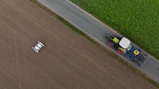 Aus der Vogelperspektive: Ein Roboter auf einem Feld, ein Weg, auf dem ein Traktor fährt.